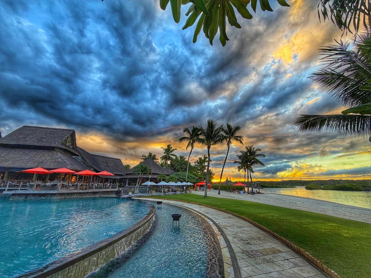 A curving turquoise pool with red parasols and thatched-roof buildings, palms silhouetted against a heavily textured blue and gold cloudscape at sunset, a lawn and calm lagoon to the right.