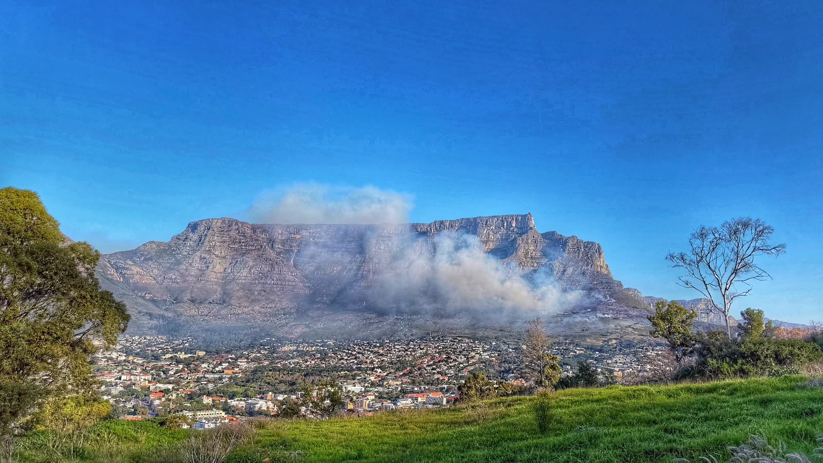 The full cliff face of Table Mountain with a large grey-white smoke plume drifting across it from a fire on the slopes, the suburbs of the city bowl below and a deep blue sky above.