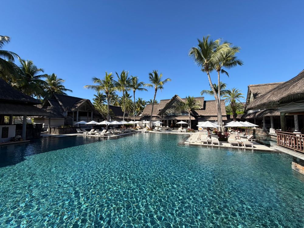 A large clear blue pool with sunloungers, white parasols and thatched-roof lodge buildings behind, tall palms against a cloudless deep blue sky.
