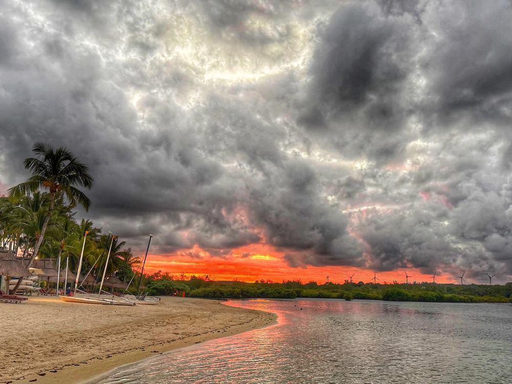 Heavy grey storm clouds above a fiery orange band of sunset, a sandy beach curving in from the left with palms, thatched huts and beached catamarans, wet sand reflecting the orange sky.