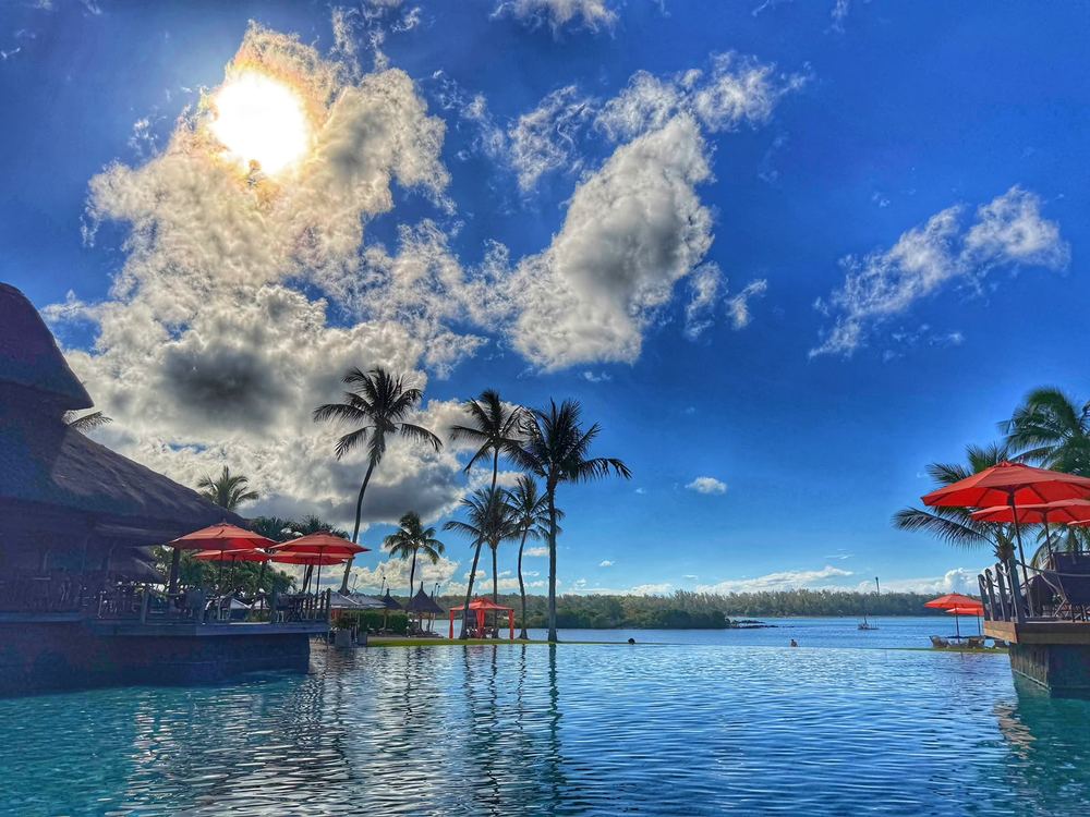 An infinity pool opening onto a lagoon with red parasols, palms and a thatched roof, the sun bursting through a large cumulus cloud in a strong blue sky.