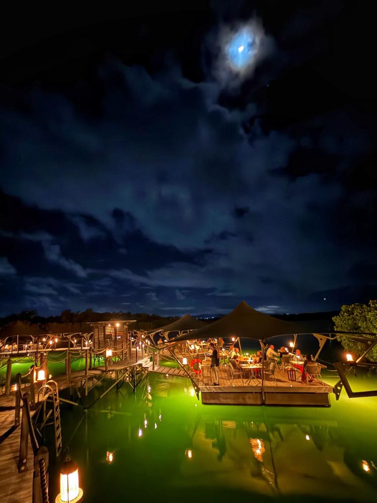 A wooden jetty restaurant built over water at night, lit by lanterns and uplights that turn the water bright green, guests at tables under a dark canopy and the moon through broken cloud.