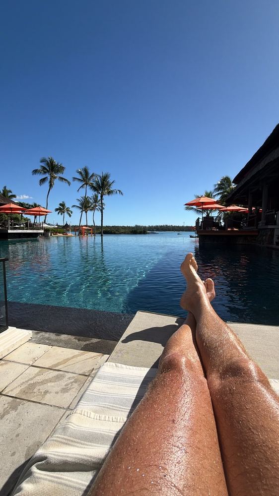 A first-person view over crossed legs on a striped sunlounger, looking across a still infinity pool to palms, orange parasols and a lagoon under a cloudless deep blue sky.