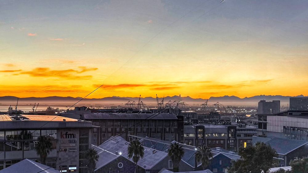 Rooftops of Cape Town's city bowl in the foreground, the working harbour with container cranes beyond, a wide orange sunrise band and distant mountains across the bay.