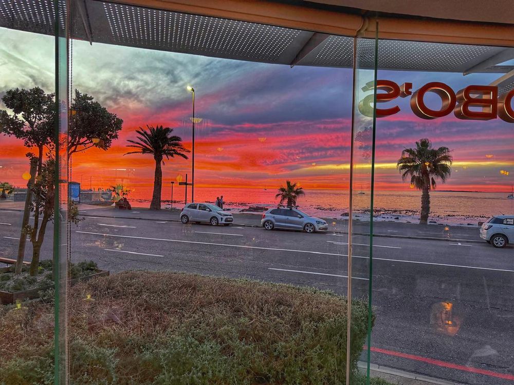 A view through a glass restaurant frontage across a coastal road to a rocky shoreline and sea, under an intense red and pink sunset with palms in silhouette.