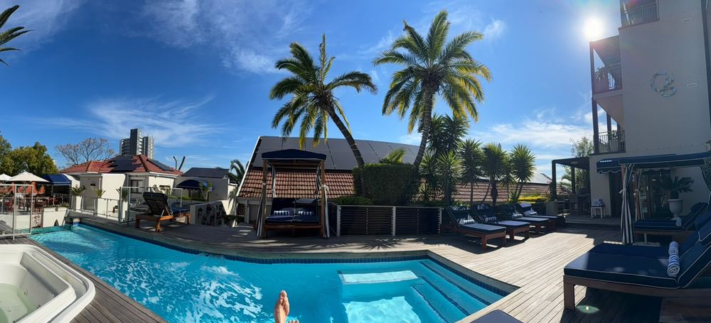 A wide panorama of a guesthouse pool with wooden decking, navy sunloungers and a canopied daybed, palms and tiled roofs behind under a clear blue sky.