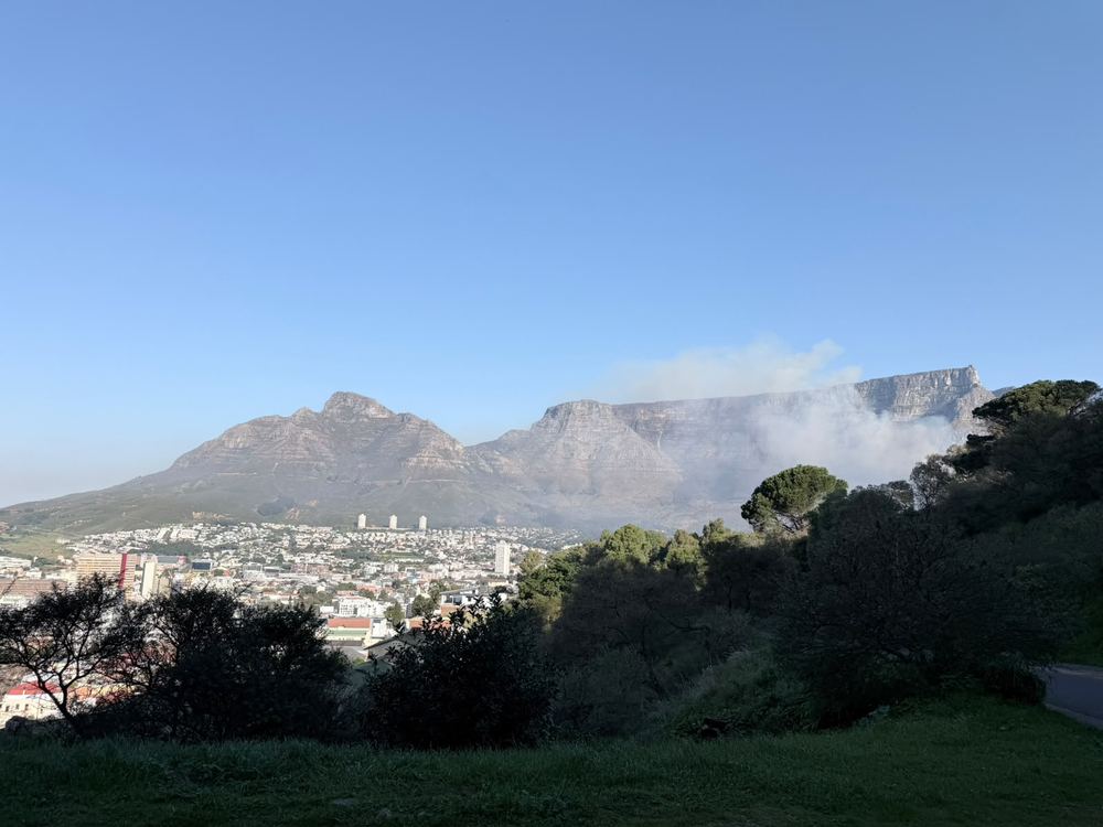 Table Mountain's flat top and the neighbouring peak seen across a green foreground slope, a plume of white smoke rising from the mountain, city suburbs along its foot.