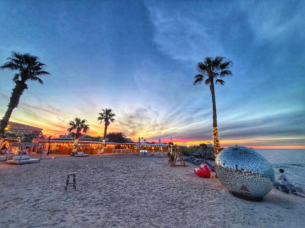 A wide sandy beach at dusk with a string-lit beach bar, illuminated LOVE letters, palms wrapped in fairy lights, a mirrored mosaic sphere and a red heart sculpture on the sand.